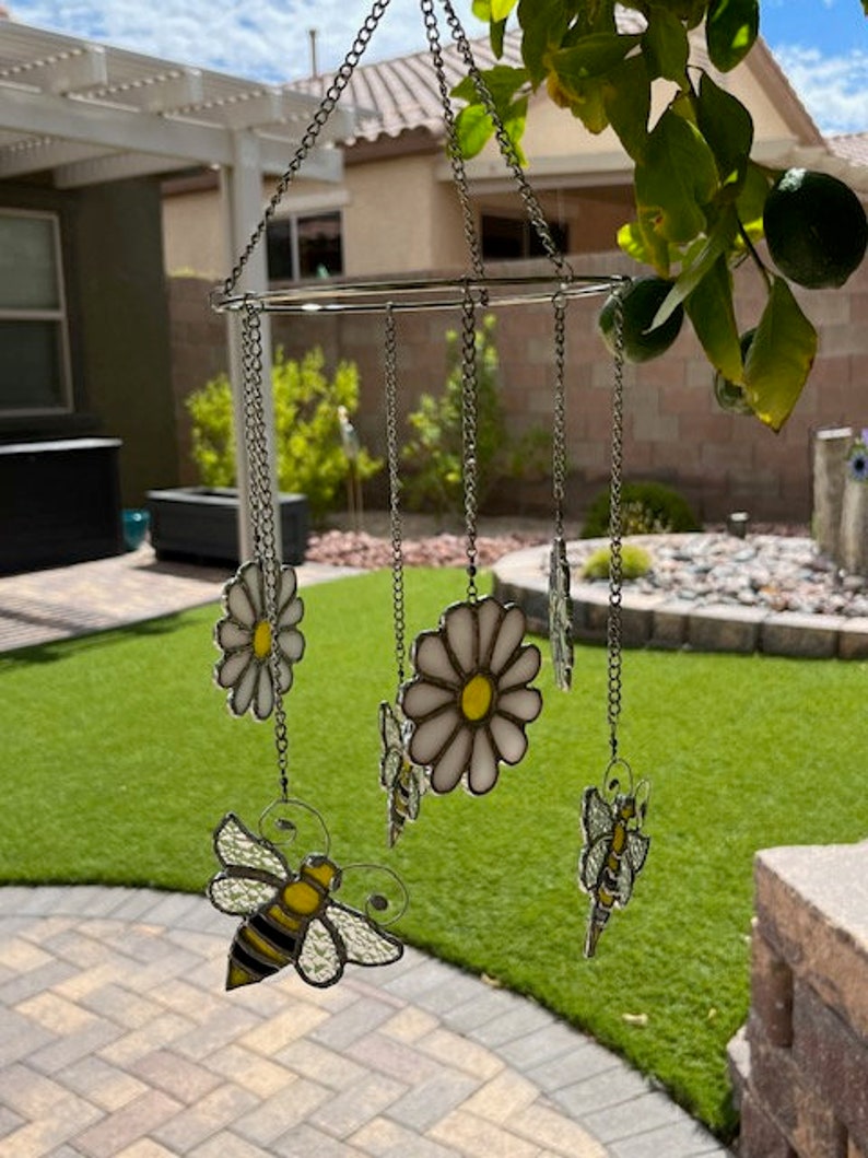 May include: A stained glass mobile featuring a white daisy, a bee, and a butterfly design. The mobile is suspended from a chain and hangs in the air.
