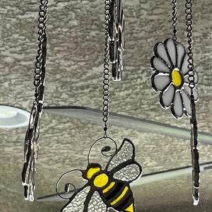 May include: A stained glass suncatcher featuring a bee, two daisies, and a chain hanging from the top.