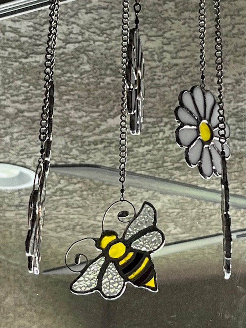 May include: A stained glass suncatcher featuring a bee, two daisies, and a chain hanging from the top.