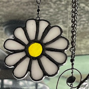 May include: A stained glass daisy suncatcher with a yellow centre and white petals. The suncatcher is hanging from a silver chain.