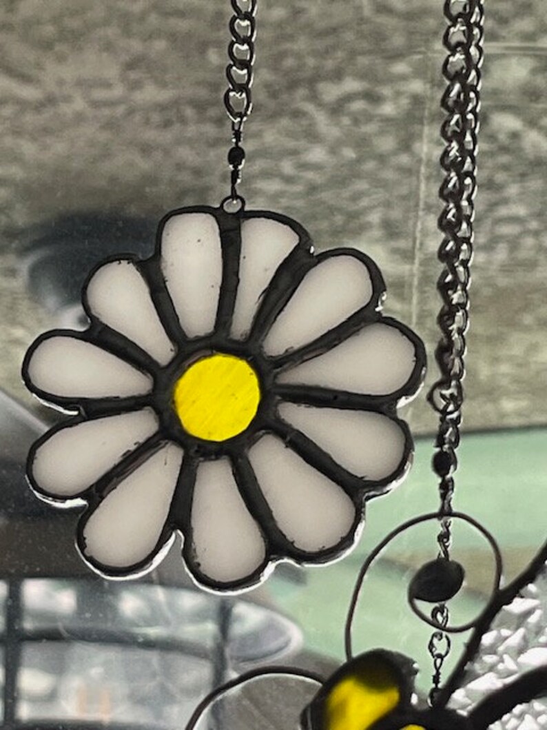 May include: A stained glass daisy suncatcher with a yellow centre and white petals. The suncatcher is hanging from a silver chain.