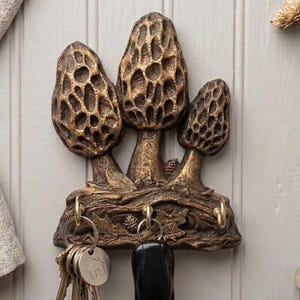 May include: A bronze-colored key holder featuring three morel mushroom sculptures. The key holder has three hooks with keys and a car key hanging from them. The key holder is mounted on a light-colored wooden wall.