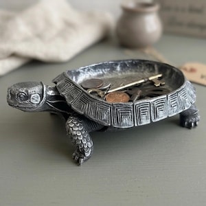 May include: A silver-colored turtle-shaped trinket dish. The dish is filled with coins and keys. The turtle has detailed features, including a textured shell and realistic head and legs. The dish is likely made of metal.