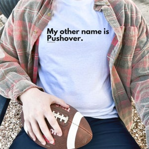 May include: A white t-shirt with the text "My other name is Pushover." printed on the front. A person is wearing the shirt and holding a brown and white football.