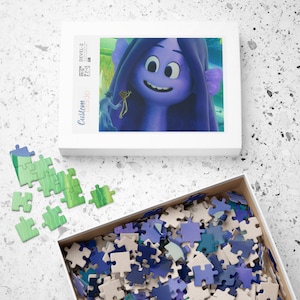 May include: A completed jigsaw puzzle with a blue and purple cartoon character with a green background. The puzzle pieces are scattered around a white box with the words "Custom Made" printed on the side.