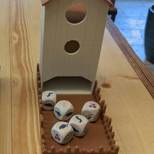 May include: A brown wooden birdhouse with a brown roof and two holes. The birdhouse is sitting on a brown wooden tray with a brown fence around the edge. There are six dice inside the tray, each with a different animal or food image on it.