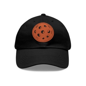 Può includere: Un cappellino da baseball nero con una grafica circolare arancione centrata che raffigura una pallina da pickleball con i suoi caratteristici fori. Il cappellino presenta una corona strutturata, una visiera curva con cuciture e un bottone.