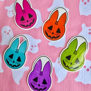 May include: Five colorful Halloween-themed stickers featuring a jack-o'-lantern bunny design. The stickers are in pink, orange, blue, green, and purple, with black facial features and a white outline. The background is pink with white ghost shapes.