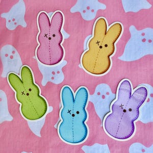 May include: Five colorful bunny stickers in pink, yellow, green, blue, and purple, each with stitched details and a stitched 'X' on the face. The stickers are on a pink background with white ghost-shaped cutouts.