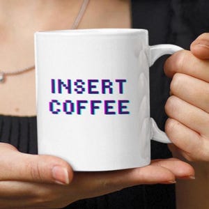 May include: White ceramic coffee mug with the text "INSERT COFFEE" in pixelated blue and purple letters. The mug has a curved handle and is held by a person.