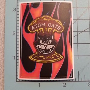 Pode incluir: Um adesivo com um gato de desenho animado usando um chap&eacute;u em forma de cogumelo com o texto "ATOM CATS" nele. O gato est&aacute; sorrindo com dentes afiados e tem um cigarro na boca. O fundo &eacute; um padr&atilde;o de chamas vermelhas, laranja e pretas.