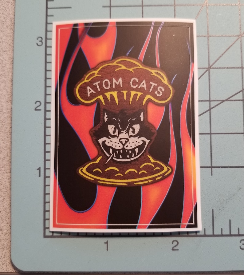 Pode incluir: Um adesivo com um gato de desenho animado usando um chap&eacute;u em forma de cogumelo com o texto "ATOM CATS" nele. O gato est&aacute; sorrindo com dentes afiados e tem um cigarro na boca. O fundo &eacute; um padr&atilde;o de chamas vermelhas, laranja e pretas.