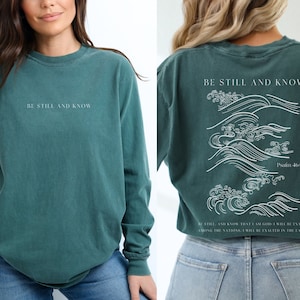 May include: Teal long-sleeve shirt with white text that reads "Be Still and Know" on the front and a white graphic of waves with the text "Be Still and Know, Psalm 46:10" on the back.