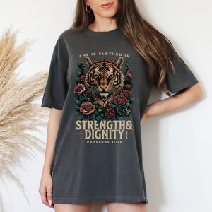May include: A charcoal grey t-shirt with a tiger illustration surrounded by roses. The text "She is clothed in strength & dignity" and "Proverbs 31:25" is printed on the shirt.