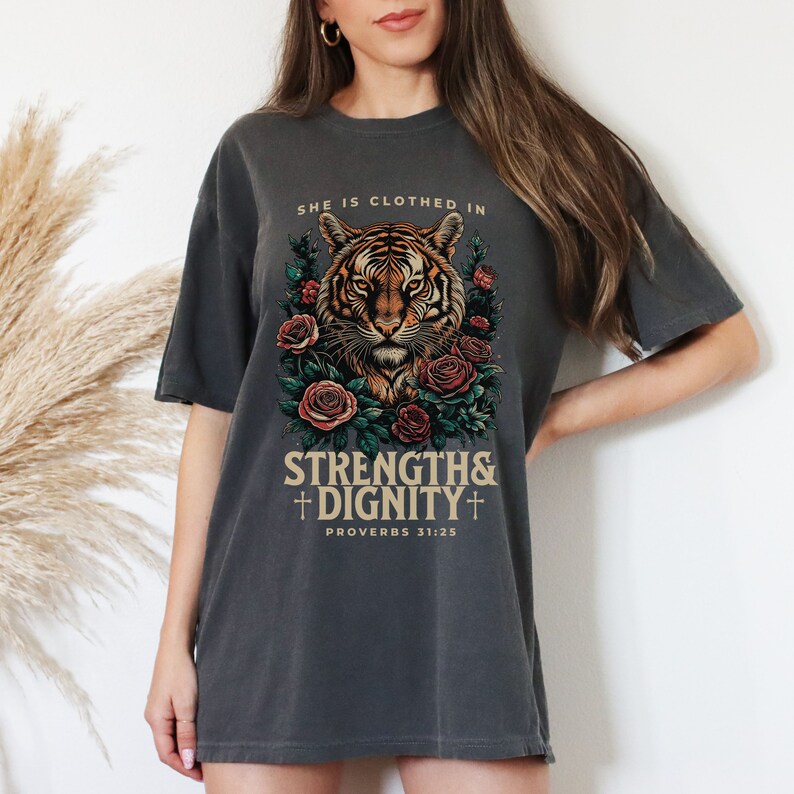 May include: A charcoal grey t-shirt with a tiger illustration surrounded by roses. The text "She is clothed in strength & dignity" and "Proverbs 31:25" is printed on the shirt.