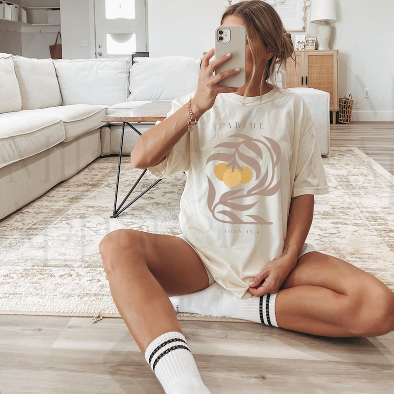 May include: A cream-colored t-shirt with a brown leafy branch design and two yellow fruit. The text "ABIDE" and "JOHN 15:4" are printed on the shirt.