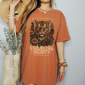 May include: Orange oversized t-shirt with a tiger graphic and floral accents. The text "She is clothed in strength & dignity" and "Proverbs 31:25" is printed on the shirt.