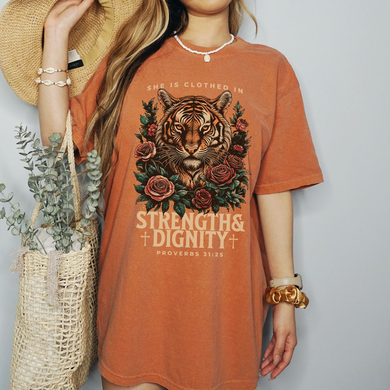May include: Orange oversized t-shirt with a tiger graphic and floral accents. The text "She is clothed in strength & dignity" and "Proverbs 31:25" is printed on the shirt.