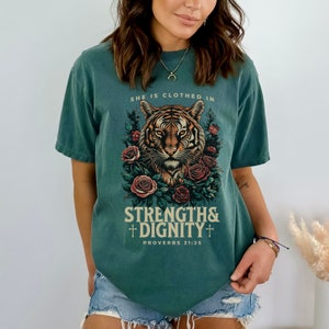 May include: A teal blue t-shirt with a graphic of a tiger surrounded by roses. The text "She is clothed in strength & dignity" and "Proverbs 31:25" is printed on the shirt.