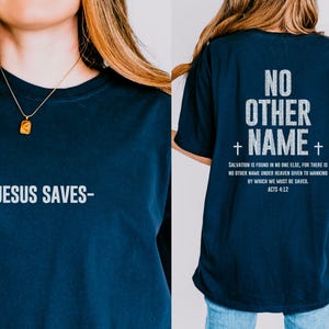 May include: A navy blue t-shirt with the text "NO OTHER NAME" in white with a cross on either side of the name. The back of the shirt has a white text quote about salvation and the scripture reference "ACTS 4:12". The front of the shirt has the text "JESUS SAVES" in white.