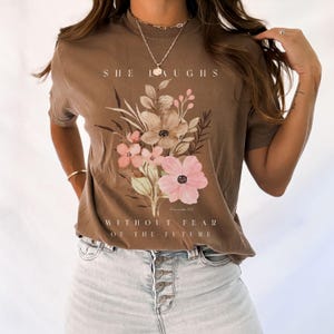 May include: A brown t-shirt with a floral design and the text "She laughs without fear of the future" and a scripture reference.