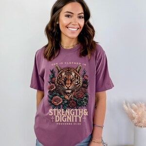 May include: Purple t-shirt with a tiger graphic and floral accents. The text "She is clothed in strength & dignity" and "Proverbs 31:25" is printed on the shirt.