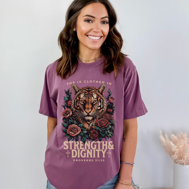 May include: Purple t-shirt with a tiger graphic and floral accents. The text "She is clothed in strength & dignity" and "Proverbs 31:25" is printed on the shirt.