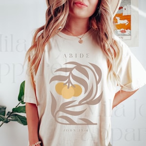 May include: A white t-shirt with a brown leafy design and yellow fruit. The text "ABIDE" is above the design and "JOHN 15:4" is below the design.