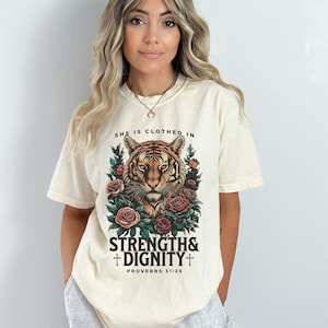 May include: A cream colored t-shirt with a tiger graphic and the text "She is clothed in strength & dignity + Proverbs 31:25"