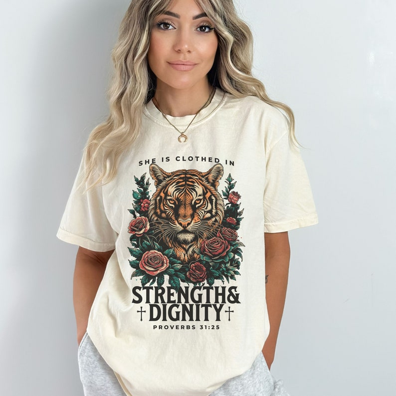 May include: A cream colored t-shirt with a tiger graphic and the text "She is clothed in strength & dignity + Proverbs 31:25"