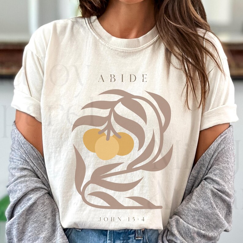 May include: A white t-shirt with a brown leafy design and three yellow circles. The text "ABIDE" is above the design and "JOHN 15:4" is below the design.