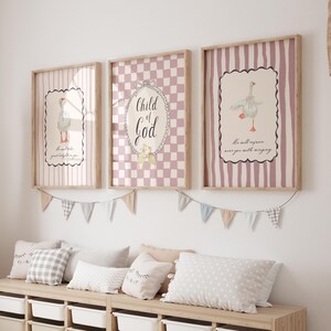 May include: Three framed prints with a nursery theme. Each print has a light wood frame. The prints feature a goose illustration, a checkered pattern with the words "Child of God", and a striped pattern with a goose illustration. A garland of pennants hangs below.
