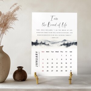 Può includere: Un calendario bianco per gennaio, sostenuto da supporti in metallo color oro, presenta il testo "Io sono il pane della vita" e un versetto biblico. Il calendario ha un'immagine di paesaggio ad acquerello e una griglia mensile.