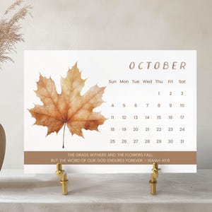 Pode incluir: Calendário de outubro decorativo com uma grande folha de bordo castanha detalhada. O calendário tem um fundo branco com uma borda castanha e um suporte dourado. O texto "Outubro" está no topo. O calendário está sobre um suporte dourado.