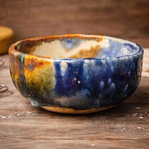 Puede incluir: Un cuenco de cerámica hecho a mano con un esmalte vibrante. El cuenco presenta una mezcla de colores, incluyendo azules profundos, naranjas y amarillos. Está sobre una superficie de madera, con herramientas de cerámica y una esponja en el fondo.