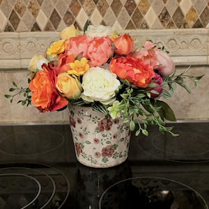 May include: A floral arrangement in a decorative ceramic pot. The bouquet features a variety of colorful flowers in shades of orange, yellow, pink, and white, with green foliage. The pot has a cream background with a floral pattern.
