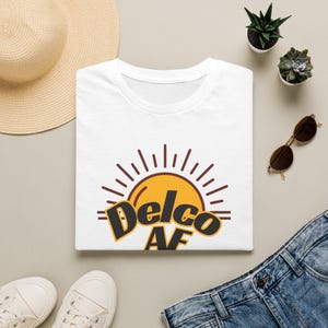 May include: A white short-sleeved t-shirt featuring a graphic of a yellow sun with brown rays and the text "Delco AF" in bold black lettering. The design is centered on the chest of the shirt, which has a classic crew neckline and short sleeves.