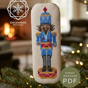 May include: A rectangular cross-stitch ornament featuring a nutcracker in a blue uniform with a red drum, yellow accents, and black boots. The design is on light fabric with a red ribbon hanger. Text includes DECEMBER STITCHES and Instant Download PDF.