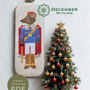 May include: A cross-stitch pattern featuring a mouse dressed as a king in a blue uniform, red cape, and gold crown. The design includes the text DECEMBER STITCHES, Instant Download PDF, and Mouse King also available in our shop. A small Christmas tree with red and gold ornaments stands beside the pattern.