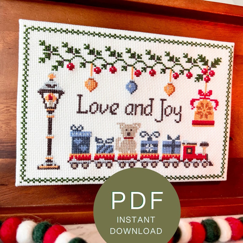Downloadable PDF for a framed cross stitch pattern with phrase Love & Joy, a Festive holiday design with a snowy street lamp, a colorful toy train, a cute dog, bells, baubles & a beautiful mistletoe.