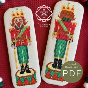 May include: Two cross-stitch patterns on off-white fabric featuring a nutcracker soldier in a red jacket, green trousers, and a gold crown. The left pattern shows the front view, and the right shows the back view. Text reads DECEMBER STITCHES.