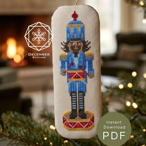 Pode incluir: Um ornamento retangular em ponto cruz sobre tecido creme com um quebra-nozes em uniforme azul, tambor vermelho, detalhes dourados e botas pretas. O design inclui uma fita vermelha e o texto DECEMBER STITCHES e INSTANT DOWNLOAD PDF.