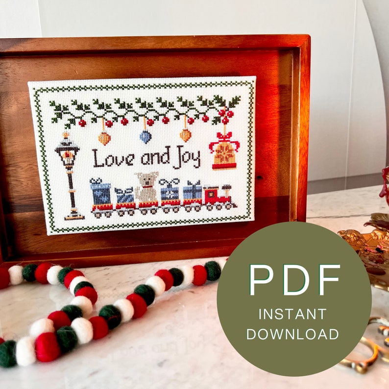 framed cross stitch pattern with the phrase Love & Joy, festive holiday design with a snowy street lamp, a toy train, cute dog, bells & mistletoe.