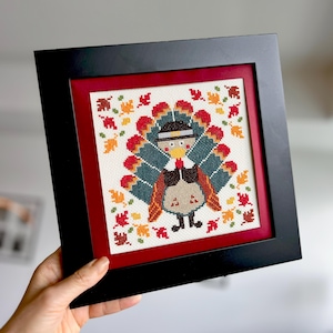 May include: Framed cross-stitch artwork depicting a Thanksgiving turkey in a pilgrim hat, with autumn leaves. The turkey has a colourful tail and is set against a cream background, framed in black with a red mount.