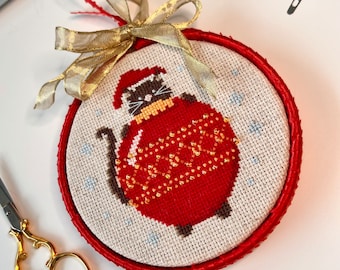 May include: A round embroidery hoop containing a cross-stitch design of a cat wearing a Santa hat and a yellow scarf, holding a large red Christmas ornament decorated with gold beads. The hoop is wrapped in red ribbon and topped with a gold bow.
