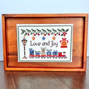 framed cross stitch pattern with the phrase Love & Joy, festive holiday design with a snowy street lamp, a toy train, cute dog, bells & mistletoe.