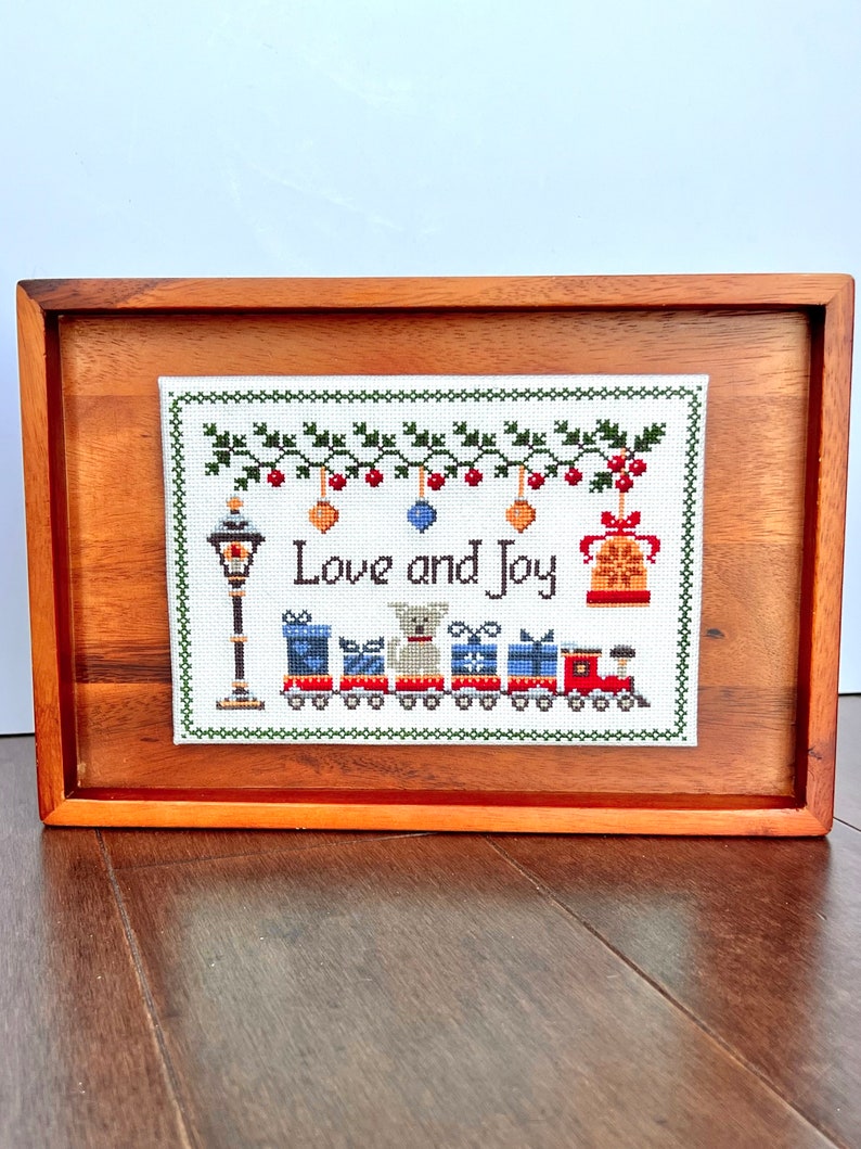 framed cross stitch pattern with the phrase Love & Joy, festive holiday design with a snowy street lamp, a toy train, cute dog, bells & mistletoe.