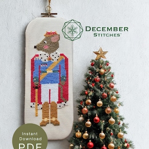 May include: A cross-stitch pattern featuring a mouse dressed as a royal figure in a blue jacket, red cape, and gold boots, with the text DECEMBER STITCHES and a snowflake logo. Beside it is a small Christmas tree decorated with red and gold ornaments.