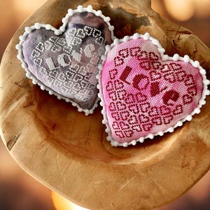 May include: Two heart-shaped decorative pillows with embroidered designs and white trim, resting in a wooden bowl. One pillow is lavender with white embroidery, the other is pink with the word "Love" embroidered in red.