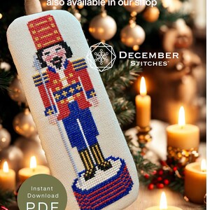 May include: A cross-stitch pattern of a nutcracker wearing a red and blue uniform with gold accents, standing on a red and blue base. The text reads "Patriotic Nutcracker" also available in our shop, DECEMBER STITCHES, and Instant Download PDF.
